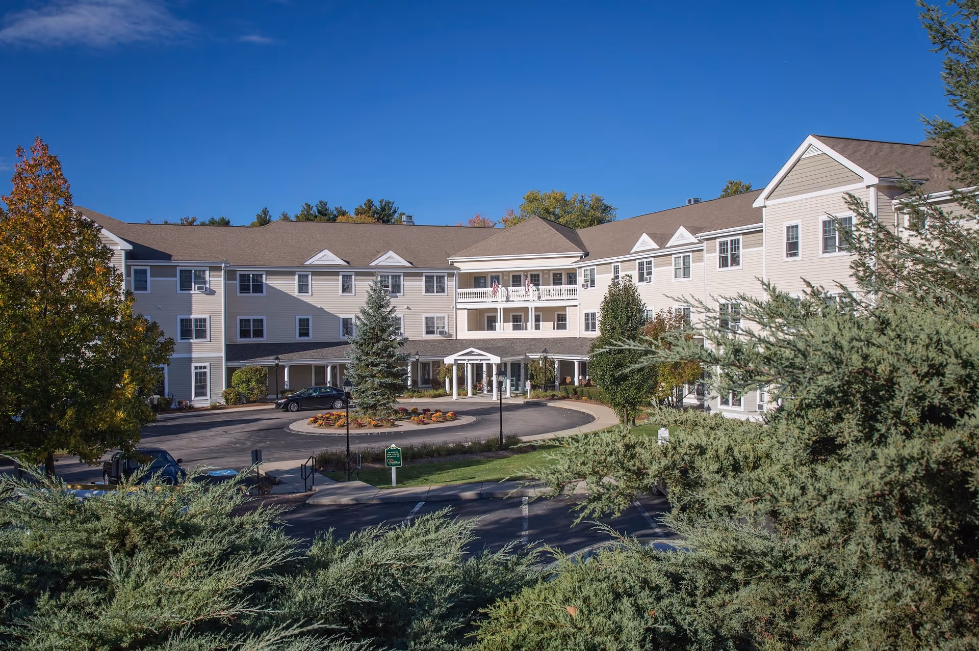 Front exterior of a three-story beige senior living building with a circular driveway, landscaping, and a clear blue sky.