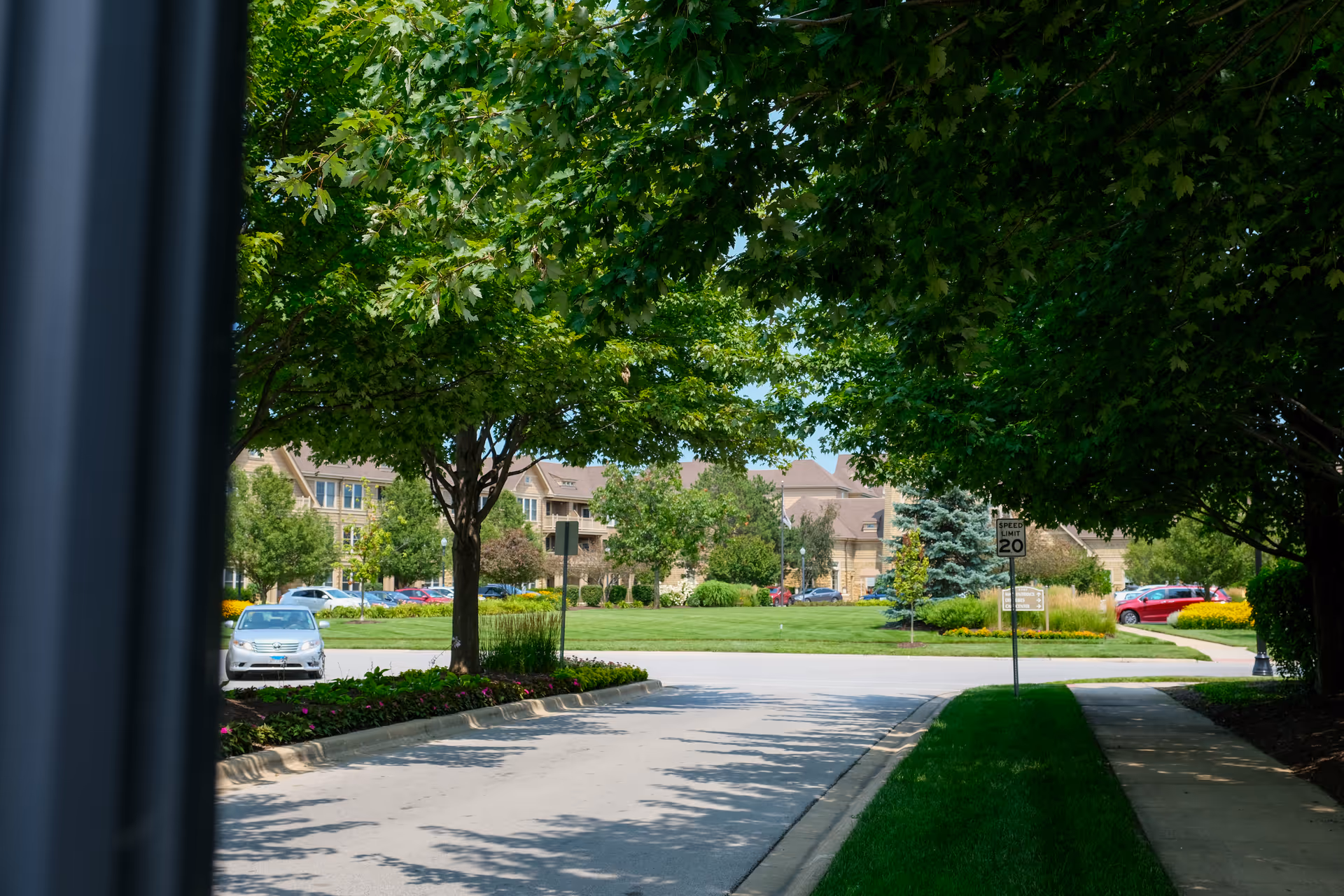 A driveway lined with green trees and flower beds leading to a grassy area with more trees and a building in the background. A speed limit sign indicating 20 mph is visible on the right side near a sidewalk.