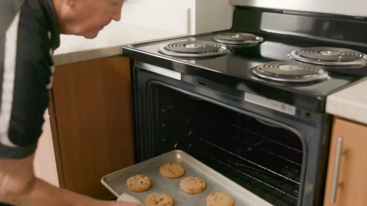 A person is removing a baking tray with freshly baked cookies from an electric oven in a kitchen setting.