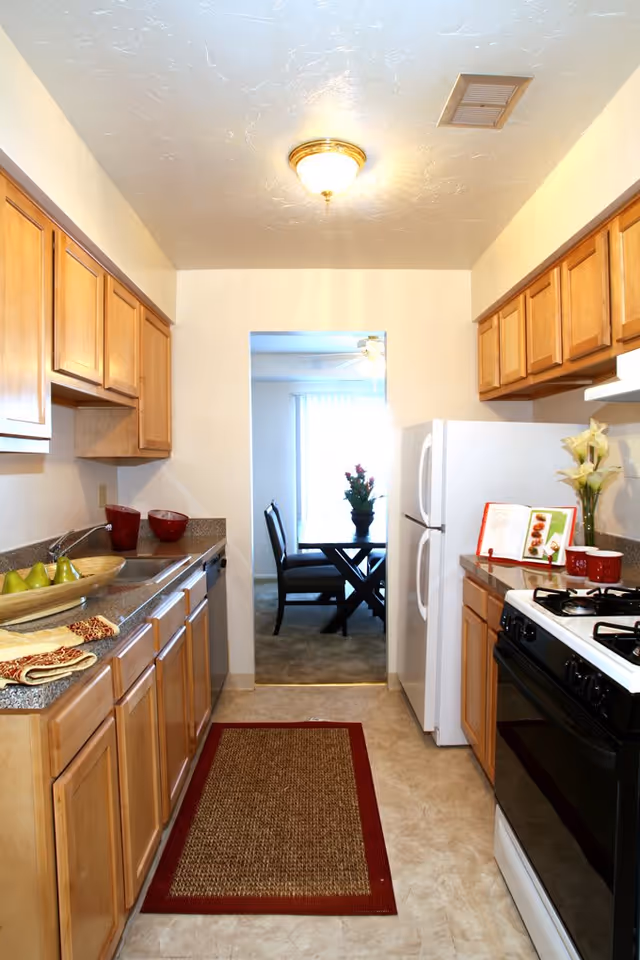 A narrow kitchen with wooden cabinets on both sides, a white refrigerator, a black stove, and a countertop with a sink and decorative items. A red-bordered rug is on the floor, and through the doorway, a dining area with a table, chairs, and a flower vase is visible.