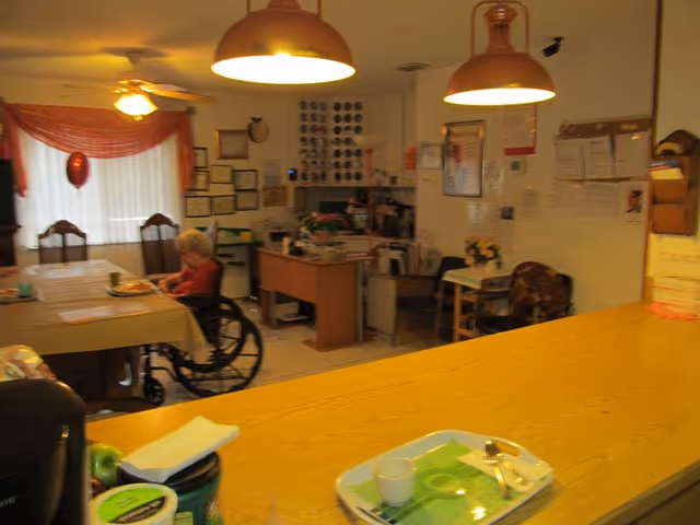 Interior view of a room in an assisted living facility with a long wooden counter in the foreground, two hanging pendant lights, a woman in a wheelchair sitting at a dining table with chairs, and a desk area with office supplies and wall decorations in the background.