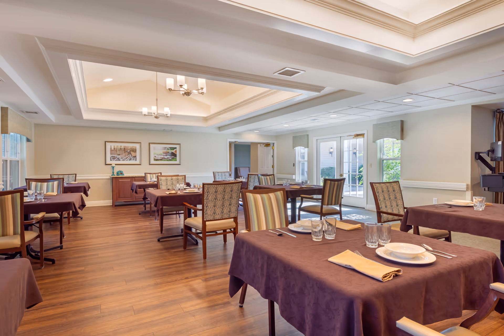 Spacious dining room with tables covered in dark tablecloths, place settings, and upholstered chairs under recessed ceiling lighting.