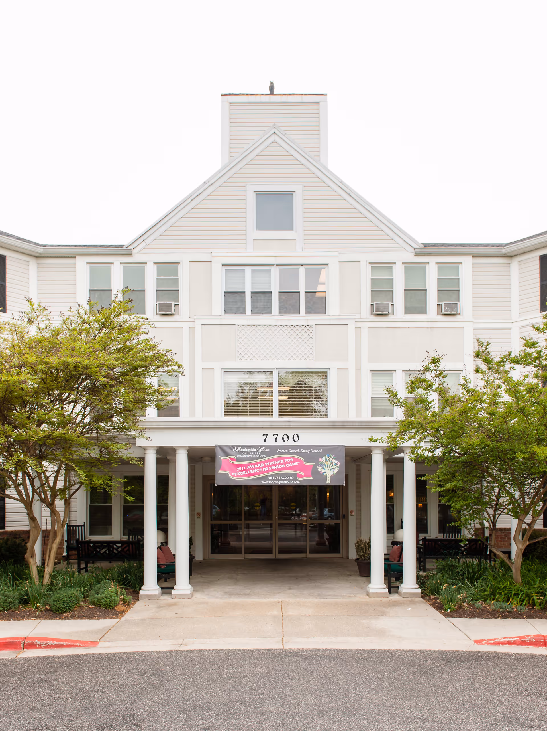 Front exterior view of a multi-story assisted living facility with white siding, multiple windows, and a covered entrance supported by white columns. There are trees and benches on either side of the entrance, and a banner above the entrance reads '2011 Award Winner for Excellence in Senior Care'.