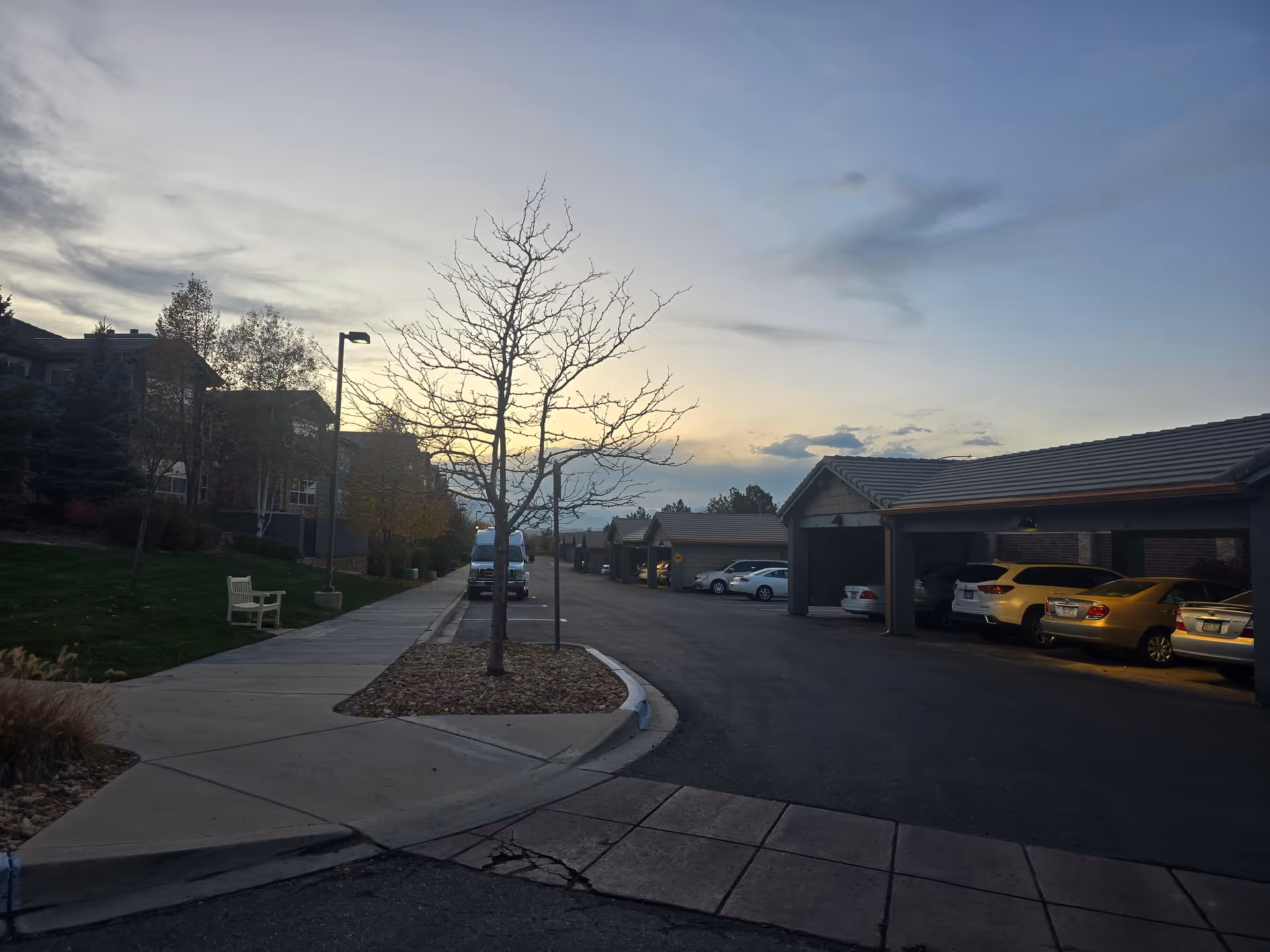 Outdoor view of a senior living facility parking area at dusk with covered parking spaces on the right, several parked cars, a sidewalk with a bench on the left, leafless trees, and residential buildings in the background under a cloudy sky.