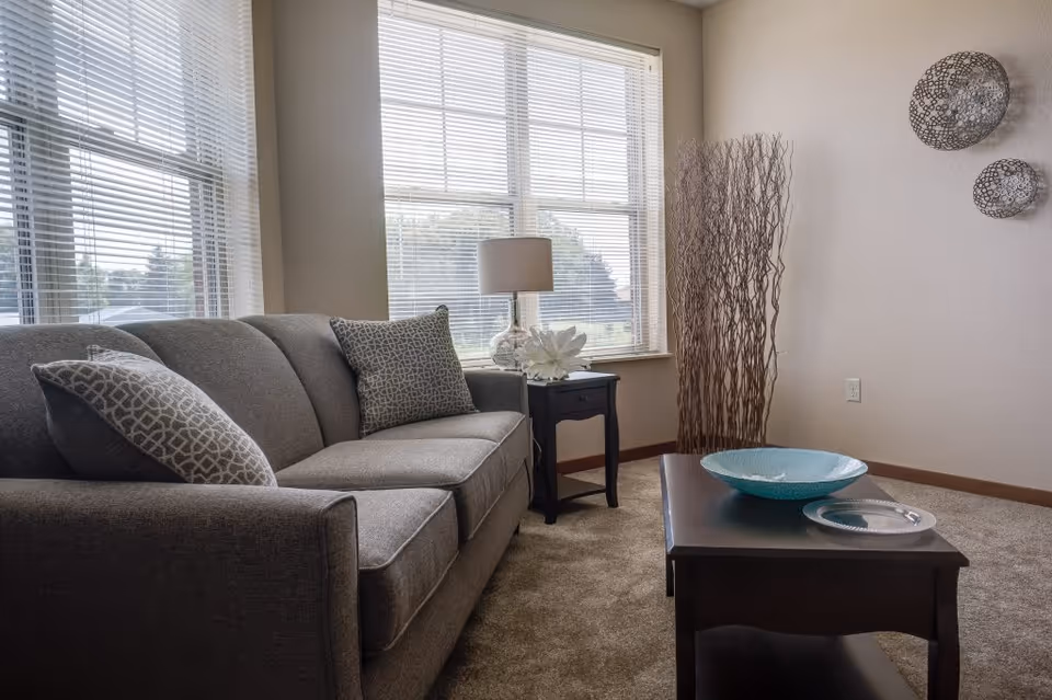 A cozy living room with a gray sofa adorned with patterned cushions, a dark wooden coffee table with decorative bowls, a side table with a lamp and a flower, large windows with blinds letting in natural light, and decorative wall art and tall dried branches in the corner.