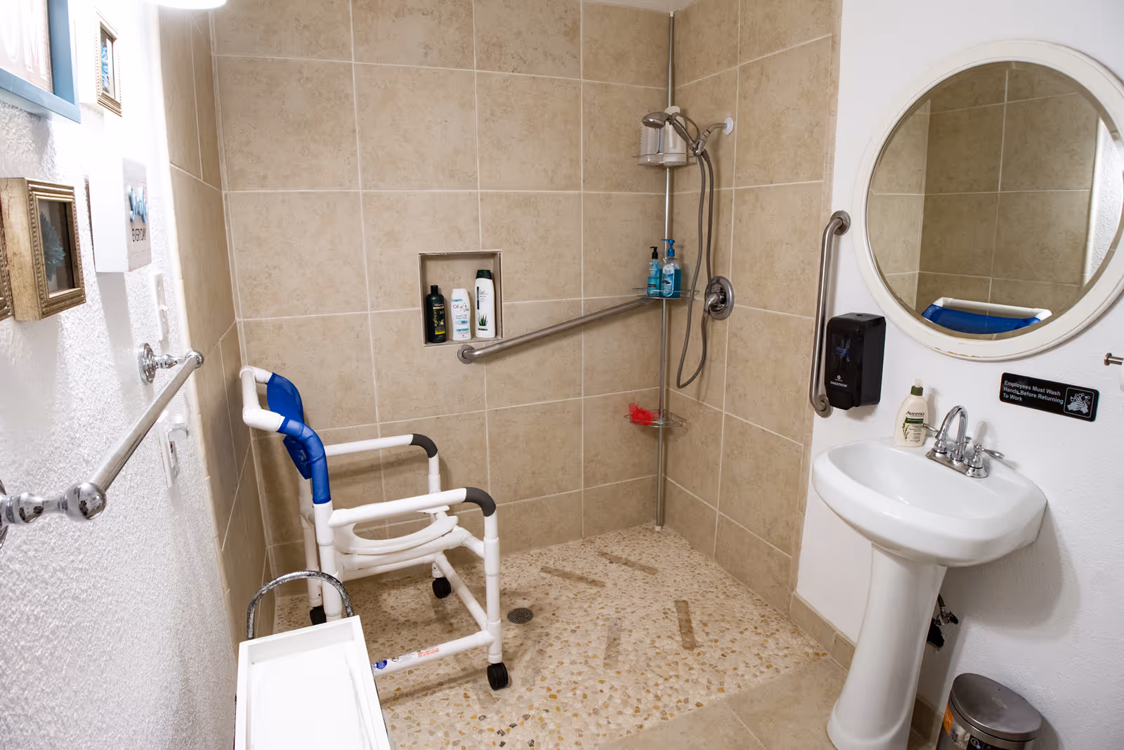 Accessible tiled bathroom with a roll-in shower, shower chair, grab bars, pedestal sink and round mirror.