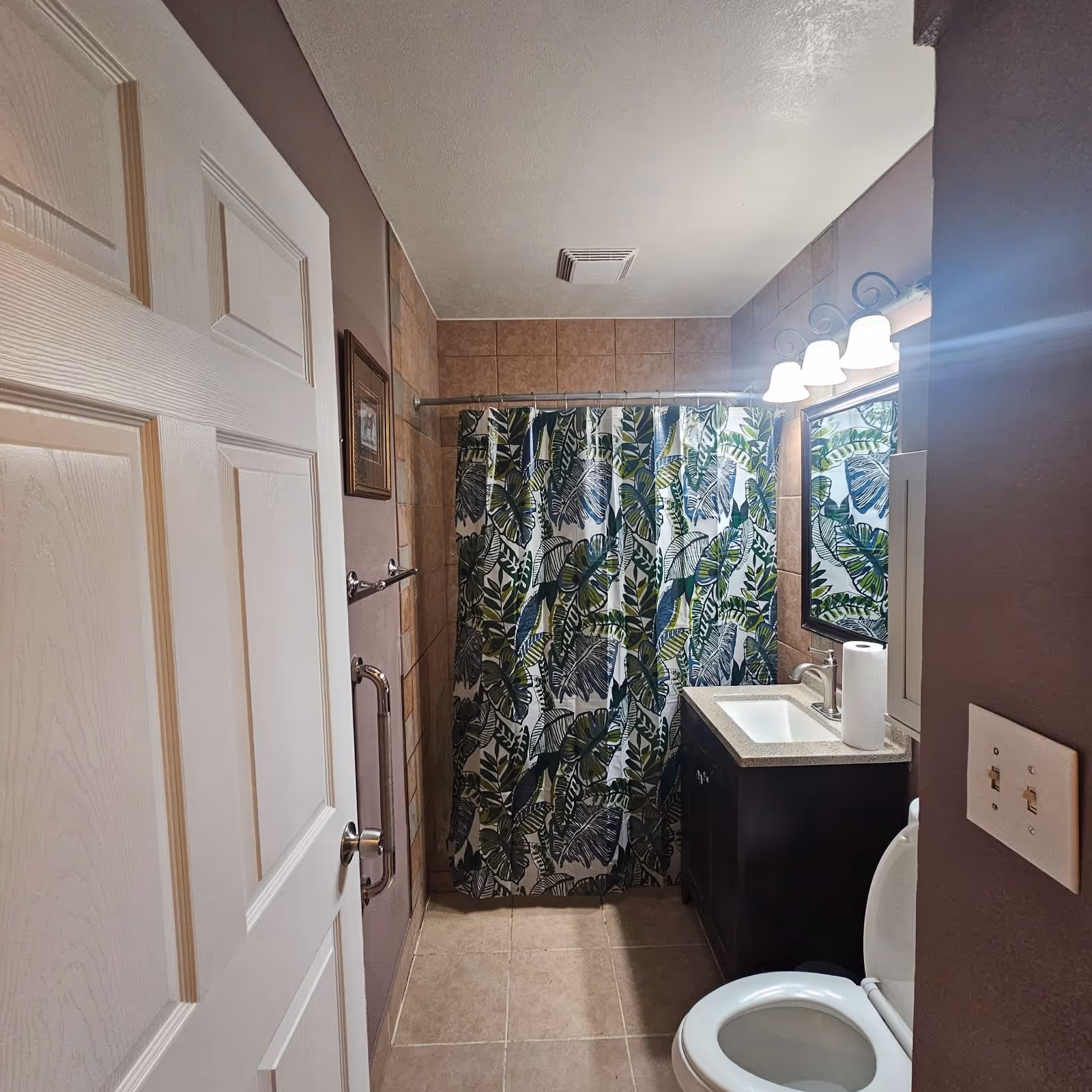 A small bathroom with a white door partially open, beige tiled walls and floor, a shower area with a curtain featuring green and blue leaf patterns, a dark wood vanity with a white sink and a paper towel roll on the counter, a mirror above the sink with three light fixtures, and a white toilet with the lid open.