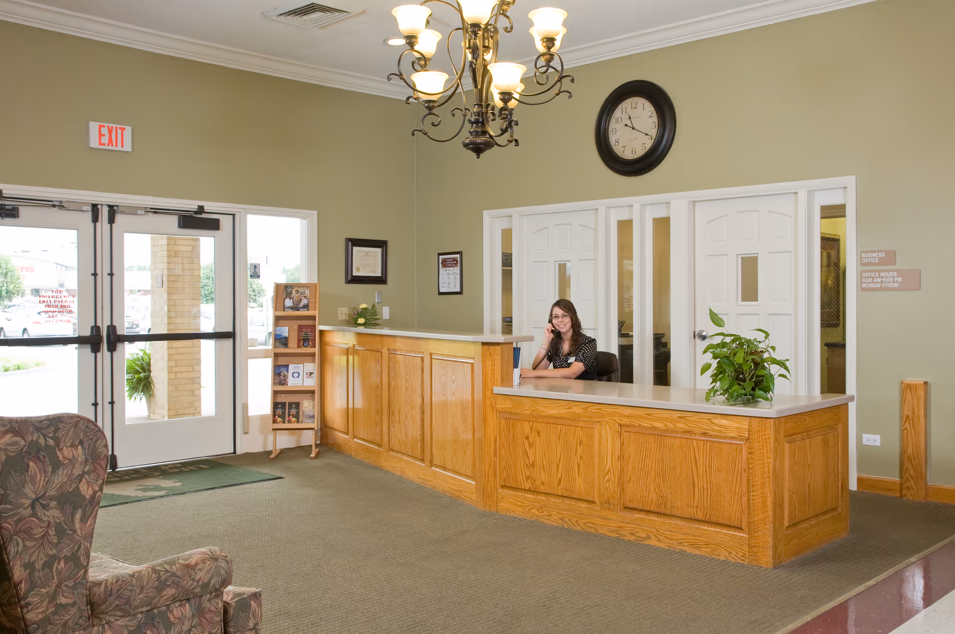 Reception area of a senior living facility with a wooden front desk, a woman sitting behind the desk on the phone, a chandelier hanging from the ceiling, a clock on the wall, and an exit door with glass panels letting in natural light.