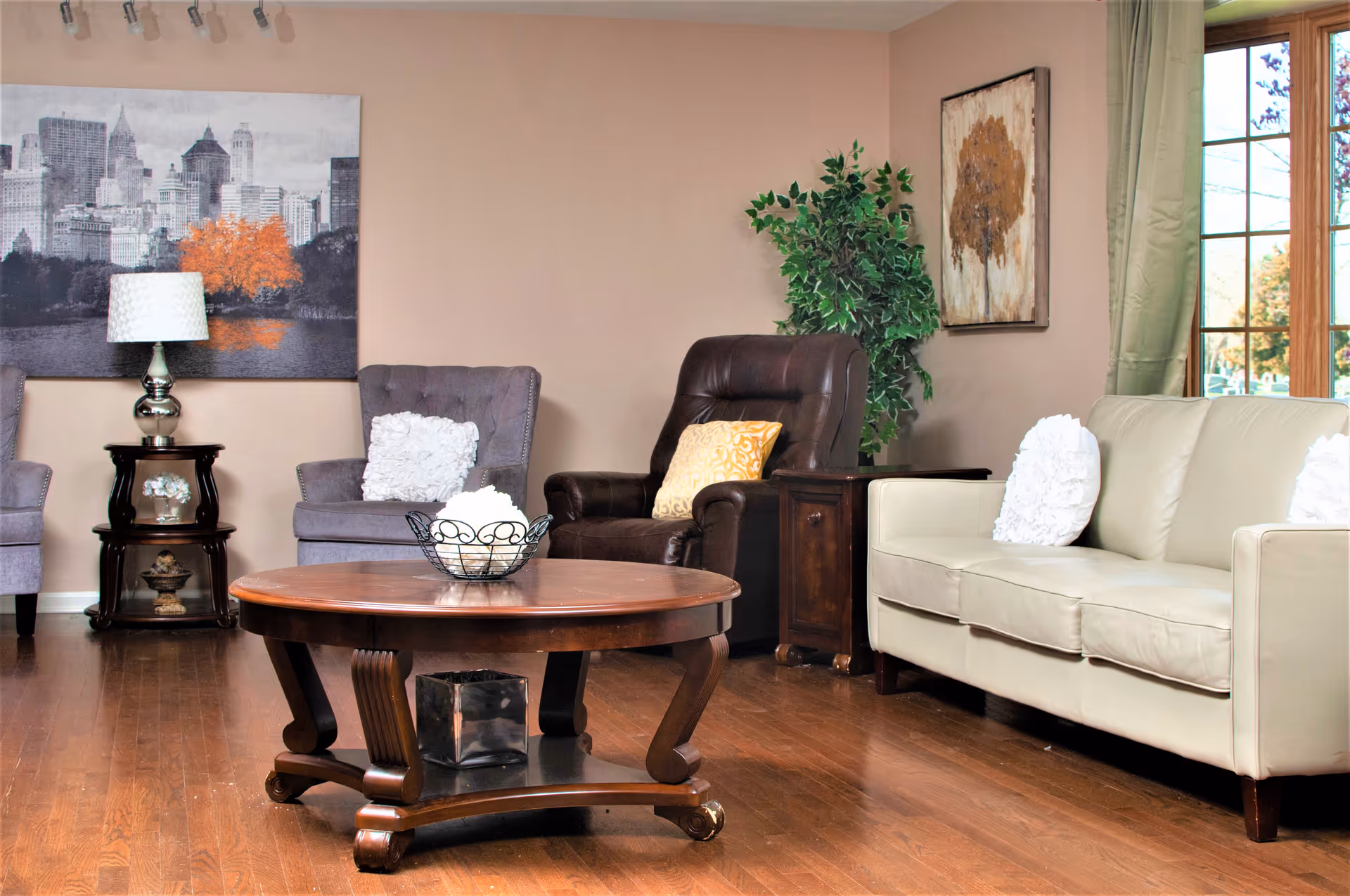 A cozy living room with a beige leather sofa, a dark brown leather armchair, and a gray fabric armchair, each with decorative pillows. A round wooden coffee table is in the center with a decorative bowl on top. The room has wooden flooring, a side table with a lamp, a large window with green curtains, and wall art including a cityscape and a tree painting. There is also a green potted plant in the corner.