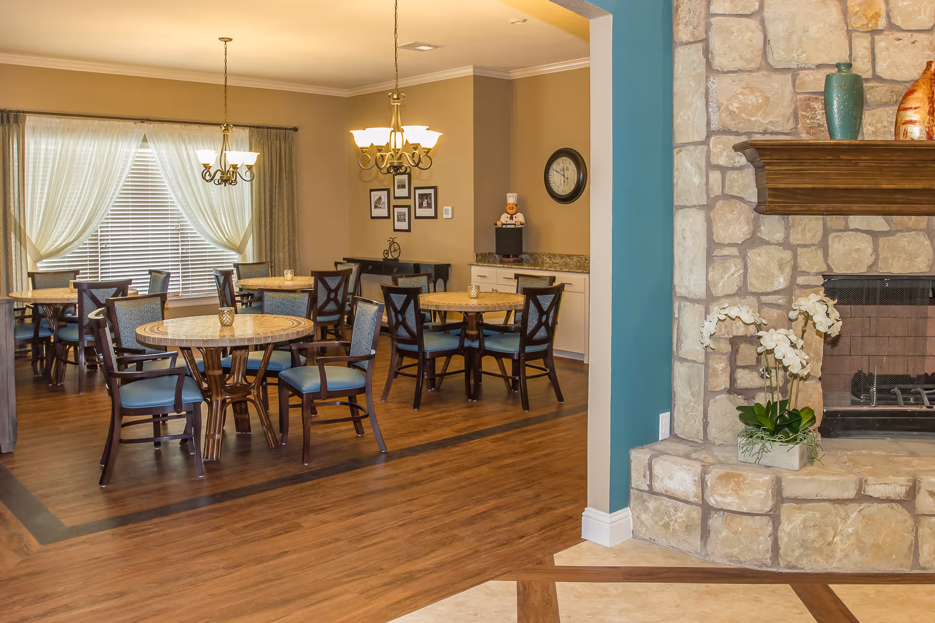 A cozy dining area with multiple round wooden tables and chairs with blue cushions. The room has warm beige walls, two chandeliers hanging from the ceiling, a large window with sheer white curtains, and a stone fireplace with a wooden mantle decorated with vases and white orchids.