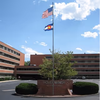Front exterior of a multi-story senior living building with flagpoles and a brick sign reading 'HOVER MANOR'.