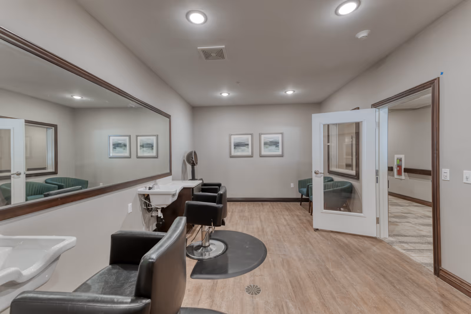 Interior view of a senior living facility hair salon area with two black salon chairs in front of sinks and a large wall mirror. The room has light-colored walls, wood flooring, two framed pictures on the far wall, and an open door leading to a hallway with carpeted flooring and a green chair.