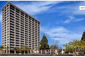 Tall multi-story residential building under a blue sky with scattered clouds, surrounded by trees and a low structure in front.