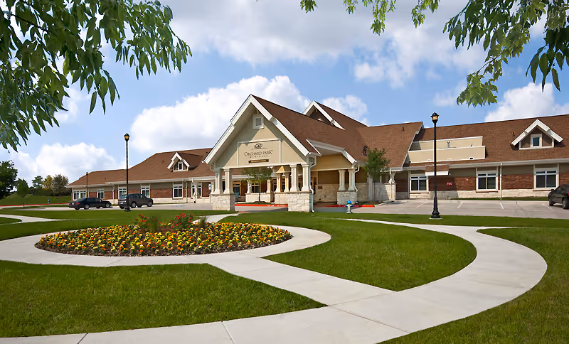 Exterior view of a senior living facility building with a large covered entrance, a circular flower bed in front, well-maintained green lawn, and a curved concrete walkway leading to the entrance. The sky is partly cloudy and there are a few cars parked near the building.