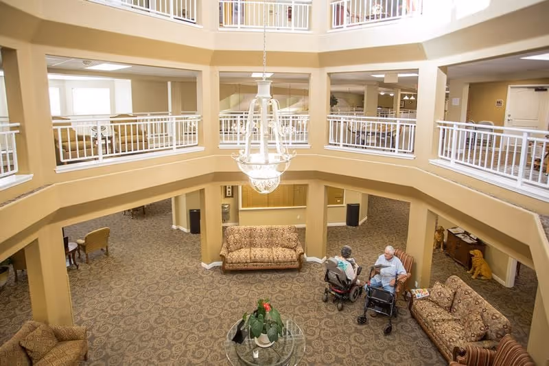 Interior view of a spacious common area in a senior living facility with patterned carpet, multiple cushioned sofas, a glass table with a plant, and two elderly individuals seated and conversing. The area is surrounded by beige walls and white railings on the upper floor, with a large chandelier hanging from the ceiling.