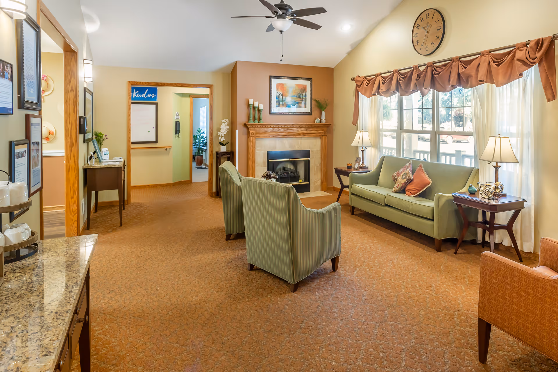 A cozy living room area in a senior living facility featuring a green sofa with decorative pillows, two green armchairs, and an orange armchair. The room has a fireplace with a wooden mantle adorned with candles and a framed painting above it. Large windows with sheer curtains and brown valances allow natural light to fill the space. There are two side tables with lamps on either side of the sofa, a ceiling fan with lights, and a carpeted floor. The walls are painted in warm beige tones, and there is a hallway visible in the background with a 'Kudos' board on the wall.
