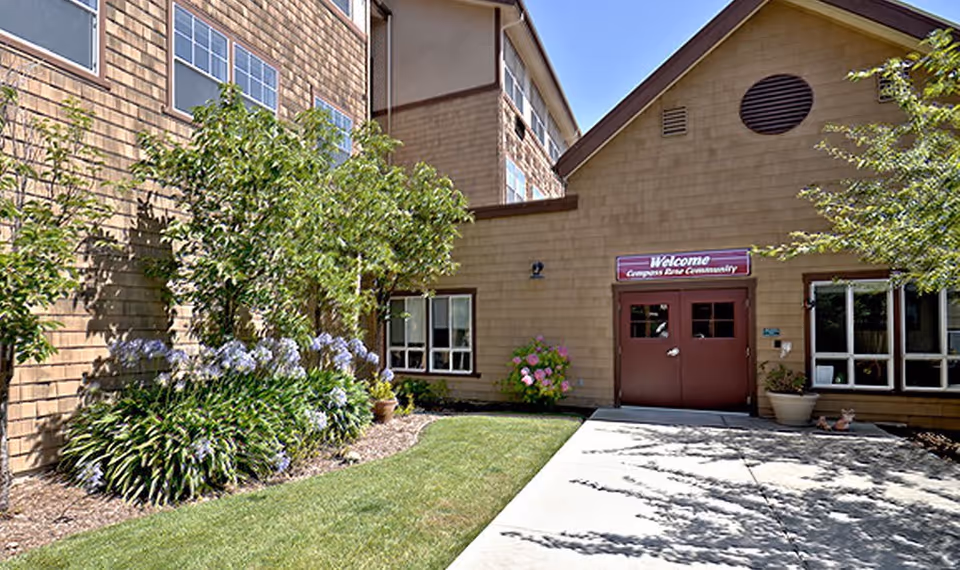 Exterior view of a building with brown siding and red double doors under a sign that reads 'Welcome Compass Rose Community'. There are windows on either side of the doors, green bushes, flowering plants, and a well-maintained lawn with a concrete walkway leading to the entrance.