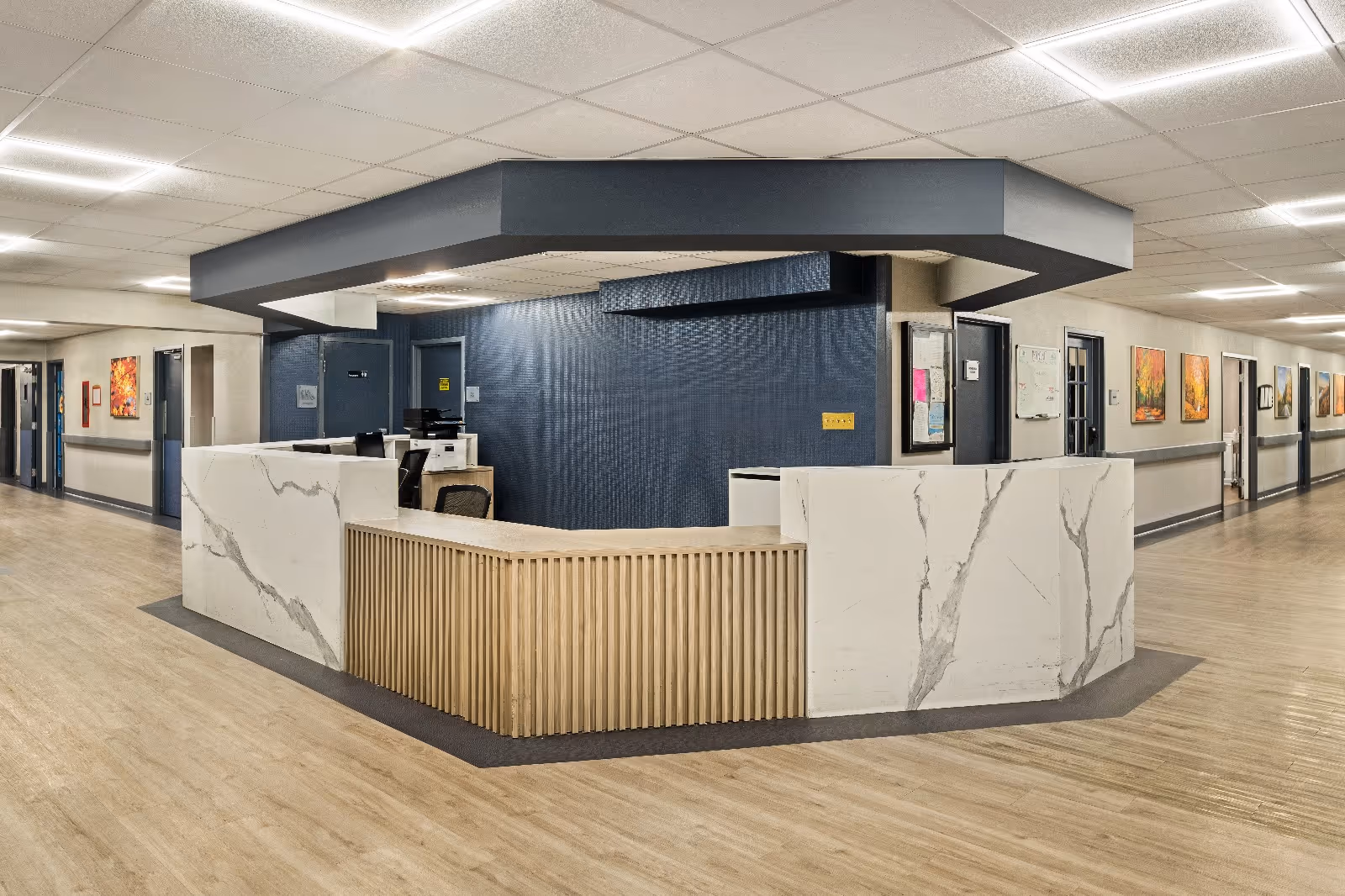 A modern reception desk area inside a senior living facility with a marble-patterned counter and wooden slats. The desk is situated in a hallway with light wood flooring, beige walls, and several doors and colorful framed paintings along the corridor. The ceiling has recessed lighting panels.
