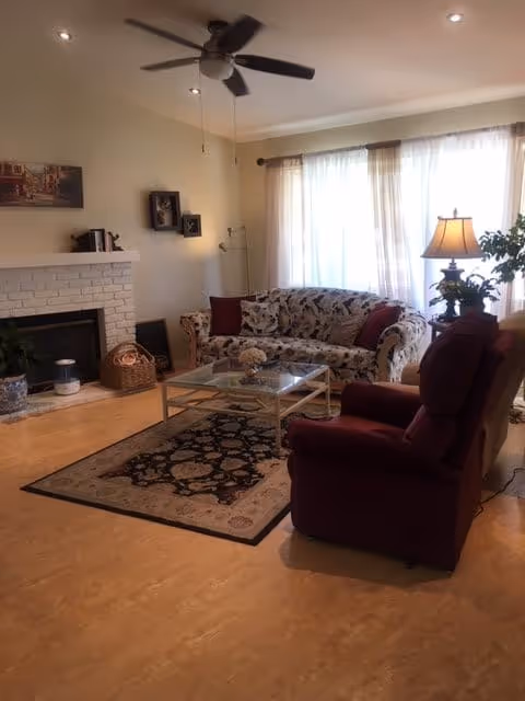 A cozy living room with a floral patterned sofa, a burgundy armchair, a glass coffee table on a decorative rug, a white brick fireplace with a basket and decor items, a ceiling fan, and a window with sheer curtains letting in natural light. A table lamp and a potted plant are placed near the window.