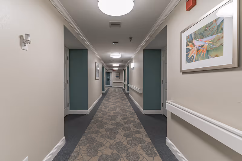 A long, well-lit hallway in a senior living facility with beige walls, teal accent panels, and patterned carpet. The hallway features white handrails on both sides, ceiling lights, framed artwork on the walls, and several closed doors along the corridor.