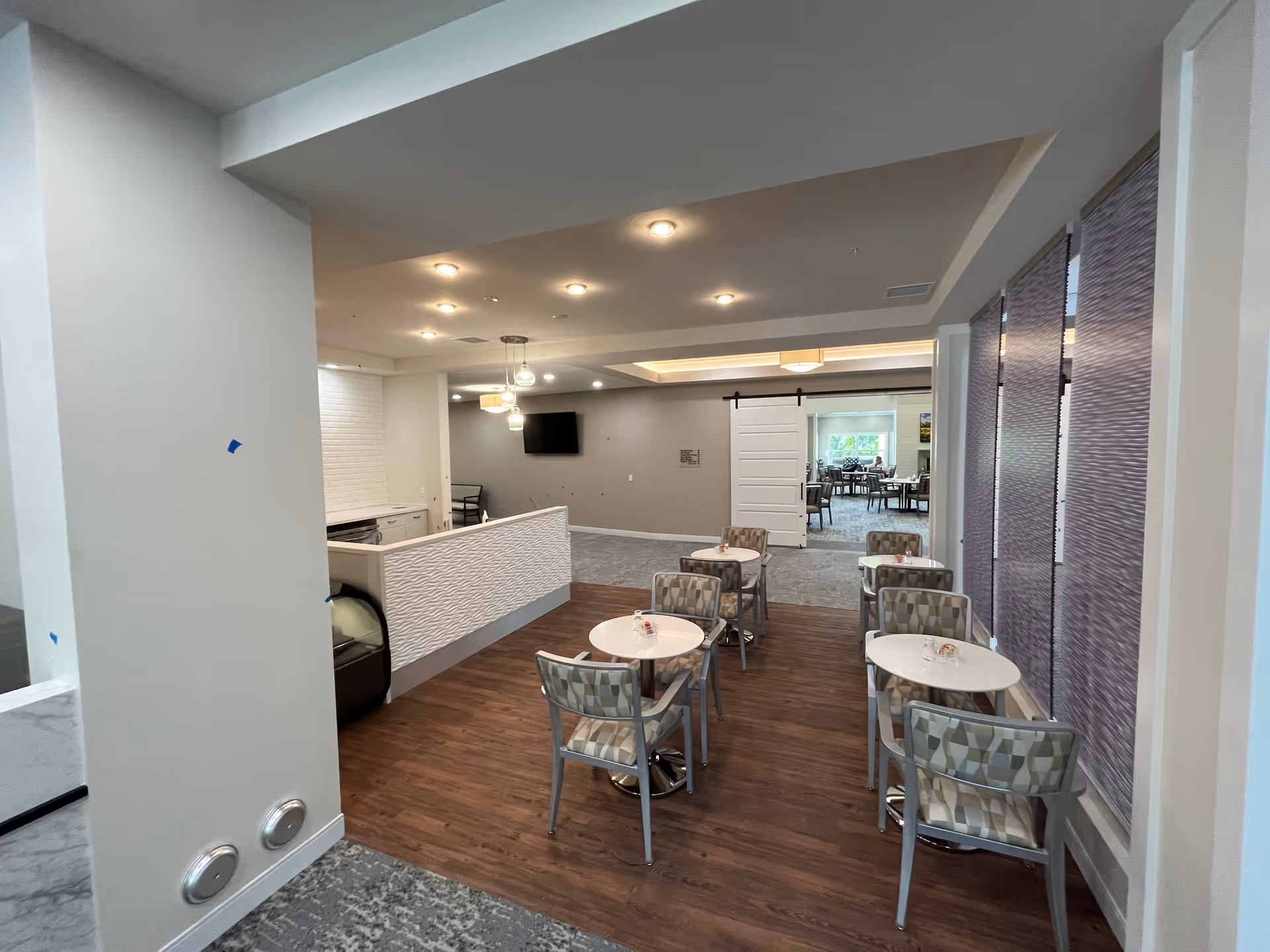 Interior view of a senior living facility dining area with small round tables and patterned chairs on a wood floor. The space includes a counter area with white textured paneling, ceiling lights, and a wall-mounted TV. In the background, there is another room with more tables and chairs visible through a wide doorway with a sliding barn door.