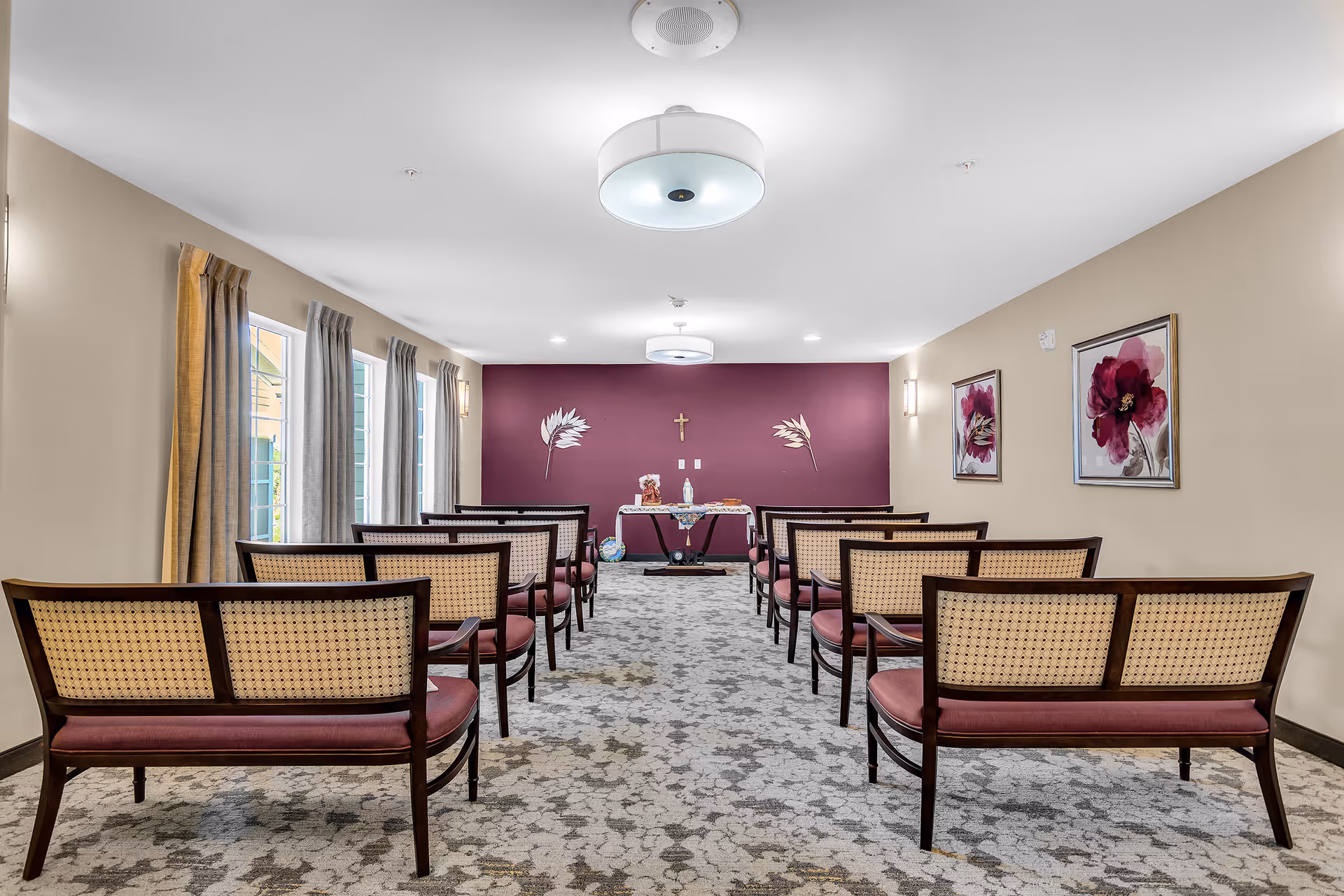Small chapel-style meeting room with rows of cane-back chairs facing a table and a cross on a purple accent wall.