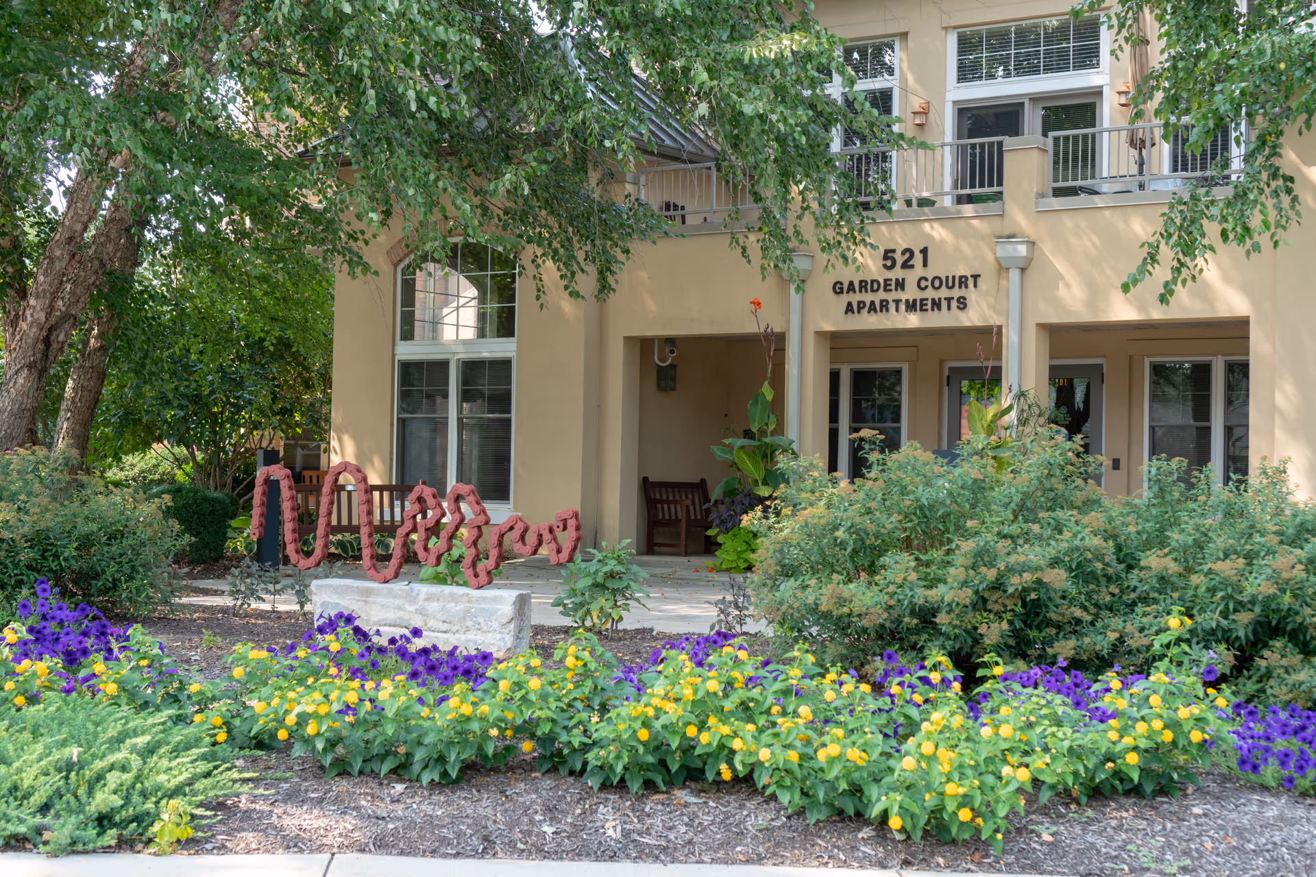 Exterior view of a beige building labeled '521 Garden Court Apartments' with a covered porch area featuring benches. The foreground has a garden with colorful flowers including purple and yellow blooms, green shrubs, and a decorative red sculpture. Trees provide shade around the building.