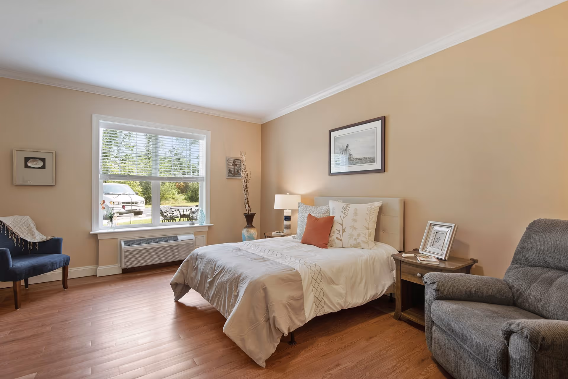A cozy bedroom with a neatly made bed featuring white and beige bedding and decorative pillows. There is a wooden nightstand with a lamp and framed photo next to the bed. A comfortable gray recliner chair is positioned nearby. The room has light beige walls, a large window with white blinds letting in natural light, and wooden flooring. A blue armchair with a throw blanket is placed near the window, and framed artwork hangs on the walls.