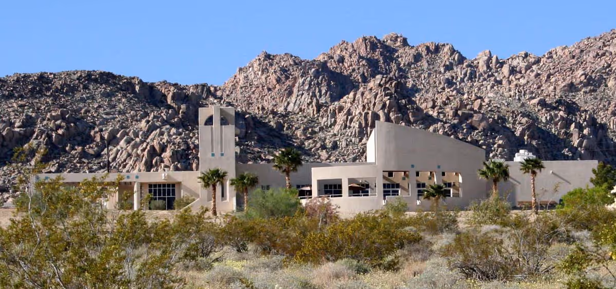 Low-slung beige building set in a desert landscape with palm trees and rocky mountains in the background.