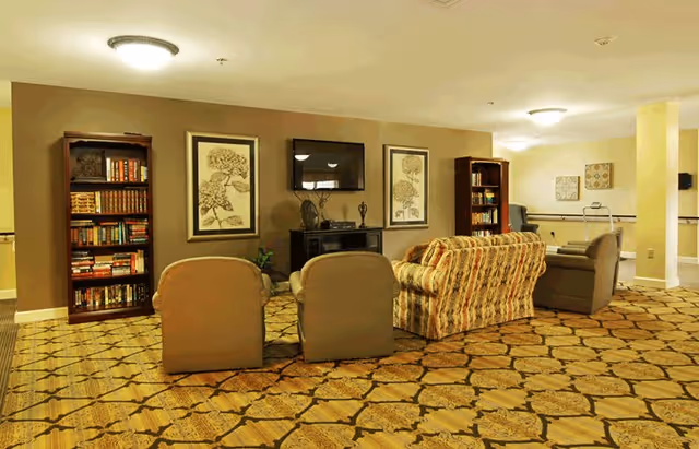 A cozy senior living facility common area with patterned carpet, two armchairs, a floral sofa, and a recliner arranged around a TV mounted on the wall. There are two bookshelves filled with books and framed artwork on the walls.