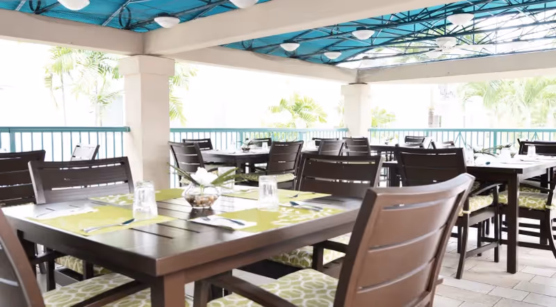 Outdoor dining area with multiple dark wooden tables and chairs with green and white patterned cushions under a blue canopy roof, surrounded by railings and tropical plants in the background.