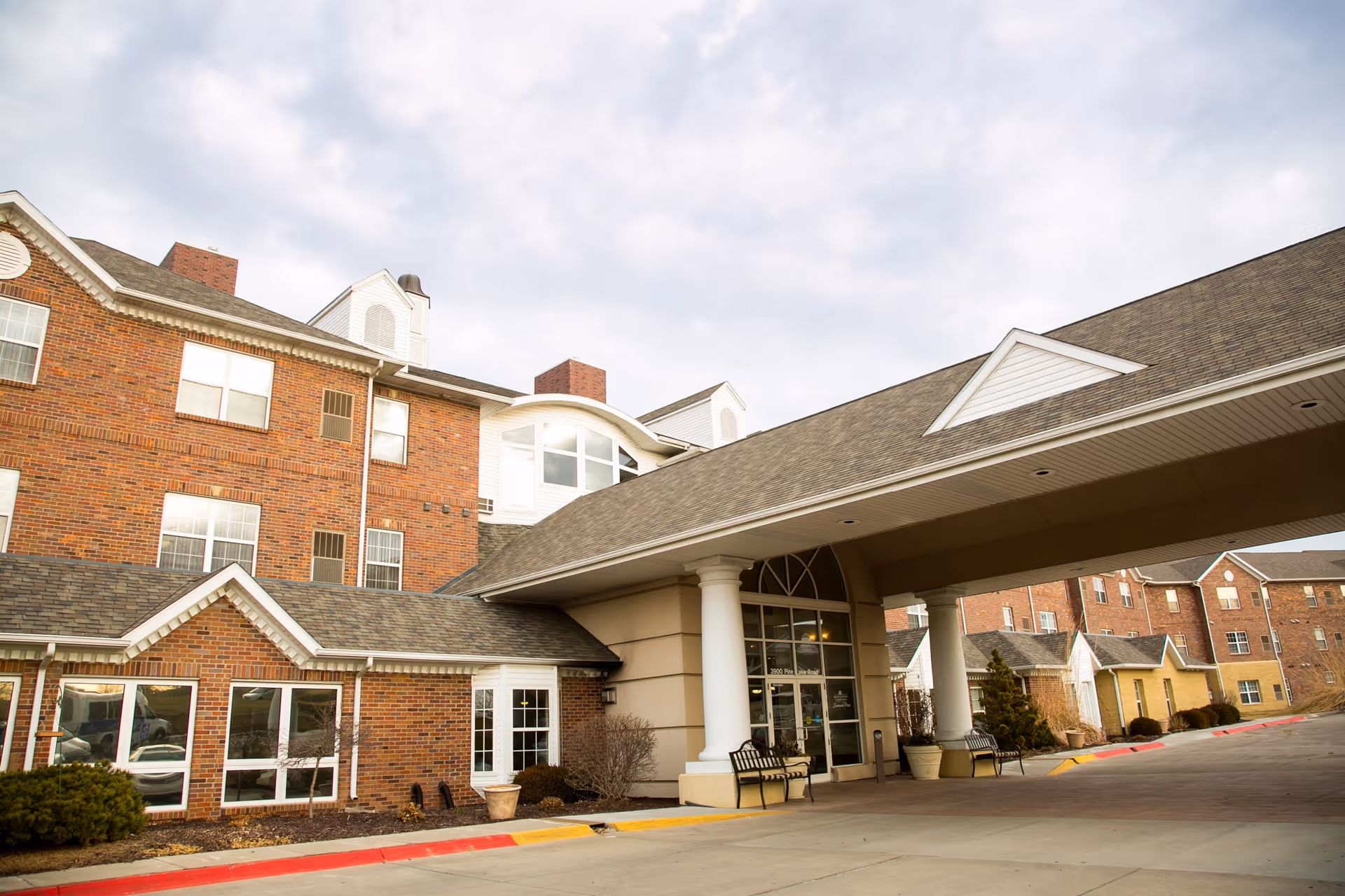 Exterior view of a multi-story brick building with white trim and a covered entrance supported by large white columns. There are benches and potted plants near the entrance, and the sky is partly cloudy.