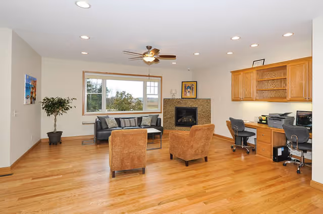 Spacious common living room with hardwood floors, a sofa and two armchairs facing a fireplace, a built-in desk area with chairs, and a large window.
