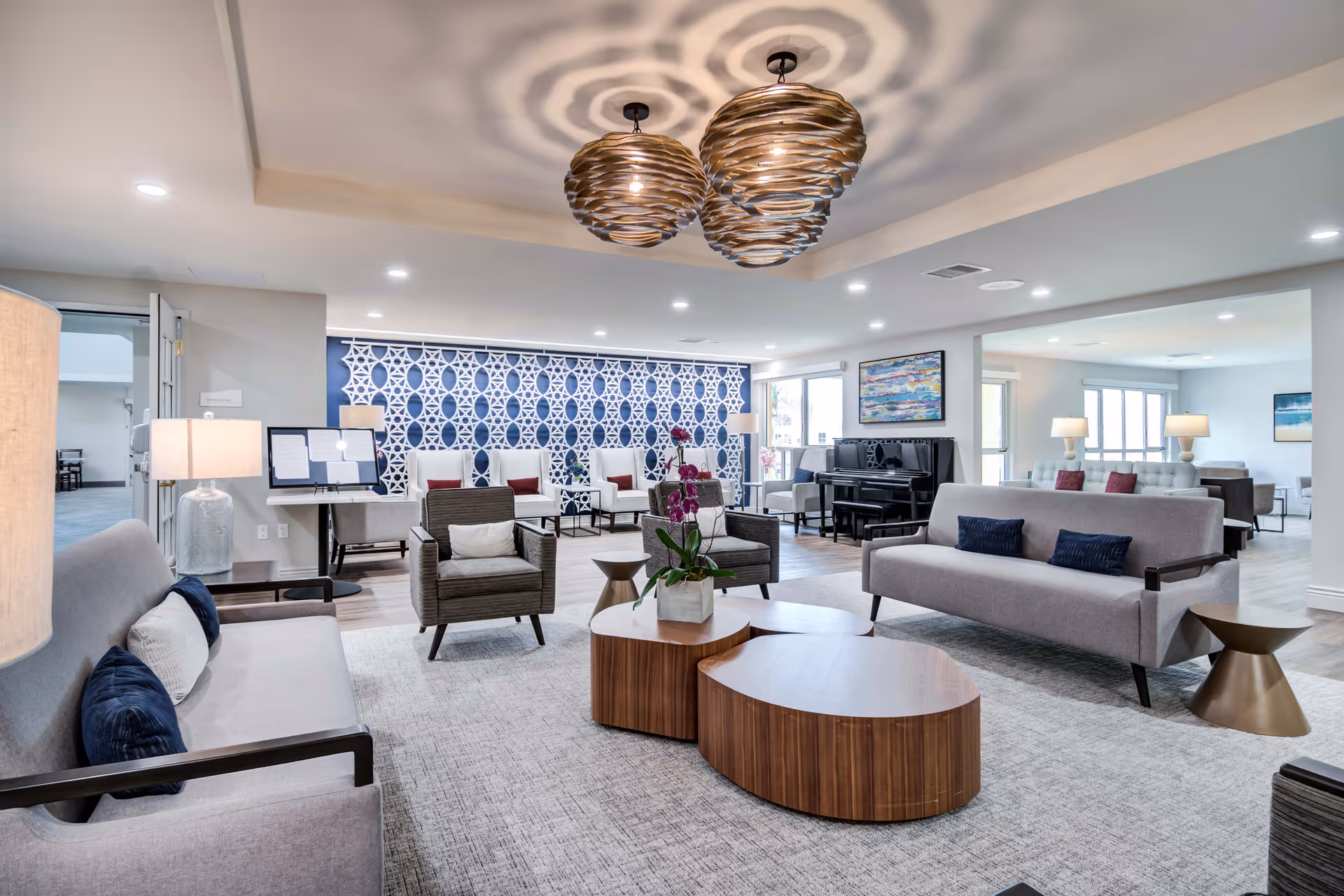 A spacious and modern living room area in a senior living facility featuring multiple gray sofas and armchairs with cushions, a wooden coffee table with a potted orchid, decorative hanging light fixtures, a patterned blue and white accent wall, a piano, and large windows allowing natural light to fill the room.