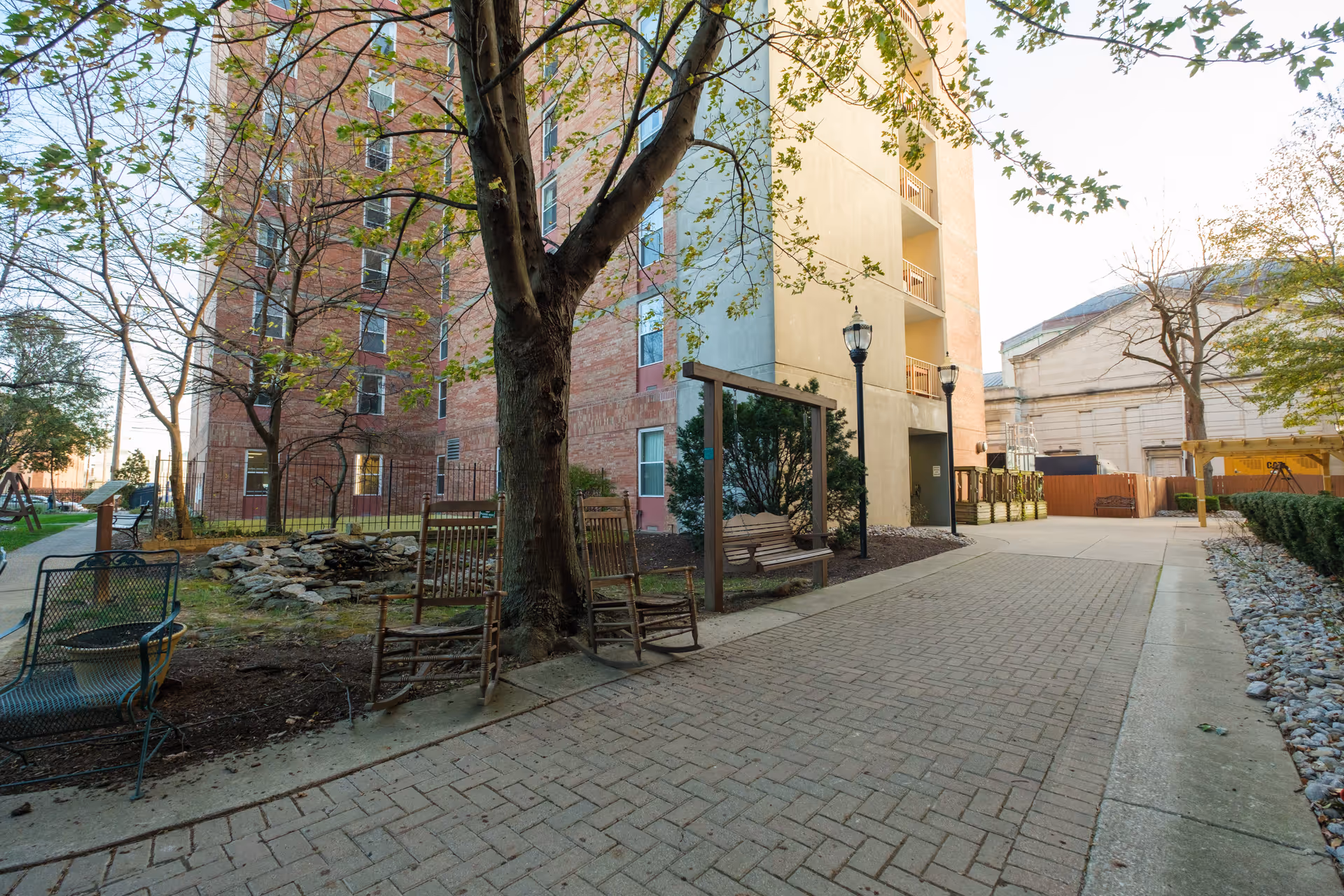 Outdoor courtyard area of Friendship House Louisville featuring a paved walkway, several trees with some leaves, two wooden rocking chairs, a wooden swing bench, a metal bench, and a small rock garden. The background shows a multi-story brick and beige building with balconies and street lamps.