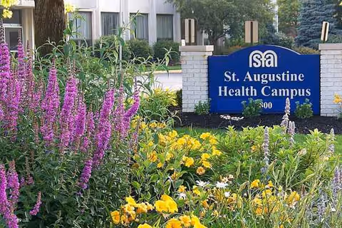 Front view of a blue St. Augustine Health Campus sign with colorful flowers.