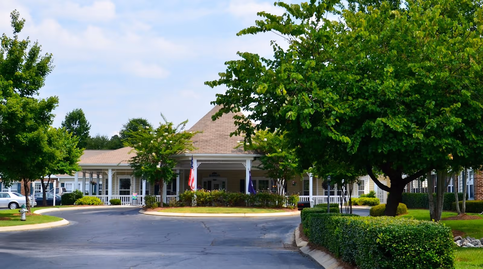 Front exterior view of Rock Hill Grove Senior Living facility with a circular driveway, green trees, bushes, and an American flag near the entrance under a covered porch.