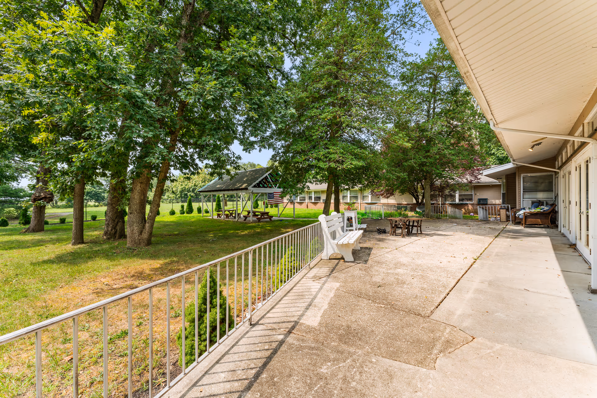 Outdoor patio area at Big Oak Rehab with white benches, a fire pit, and a covered picnic area surrounded by large trees and green grass under a clear blue sky.