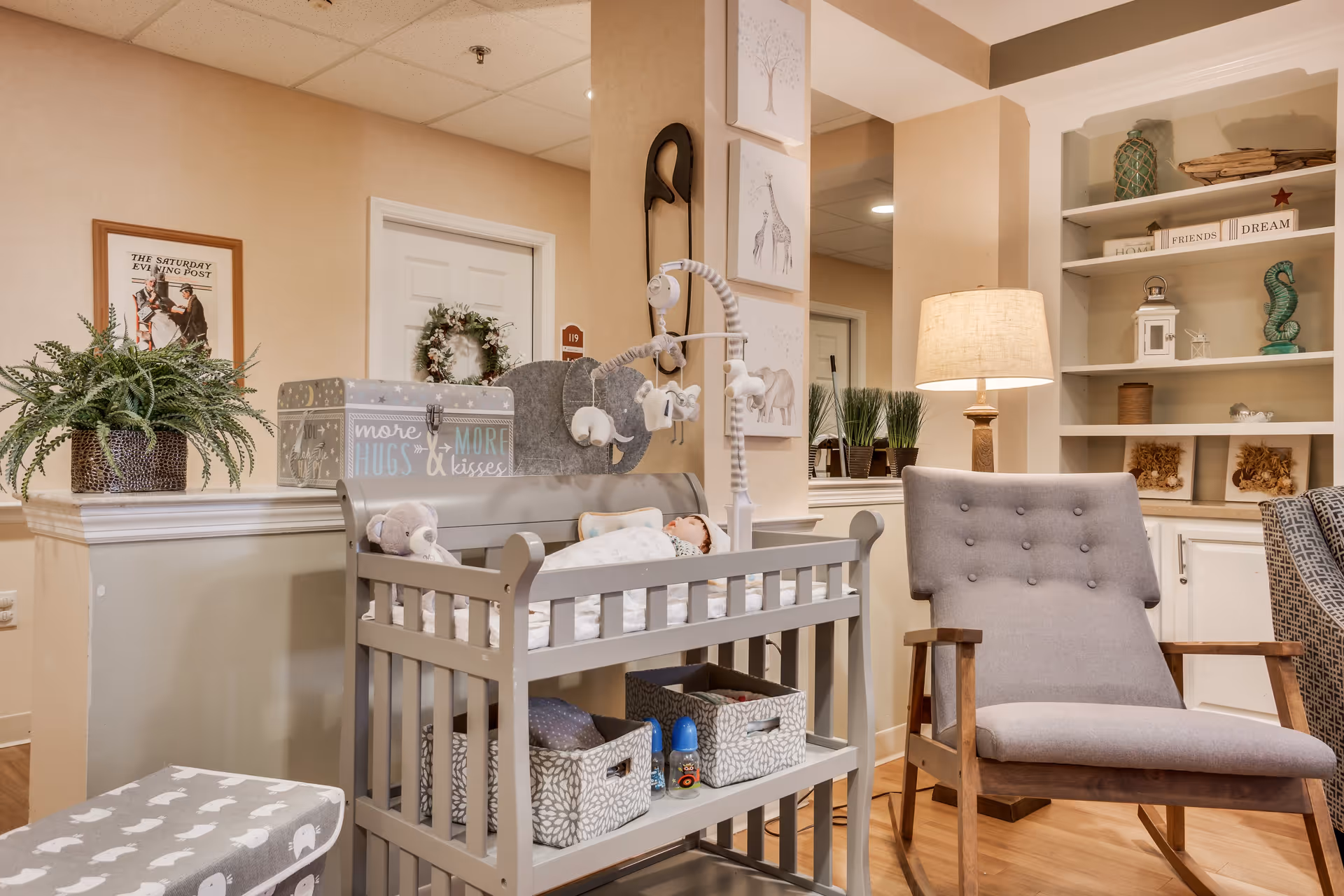 A cozy interior corner of a senior living facility featuring a gray cushioned armchair, a baby changing table with stuffed animals and storage baskets underneath, a lamp on a side table, built-in shelves with decorative items, and a framed picture on the wall. The setting has warm lighting and a welcoming atmosphere.
