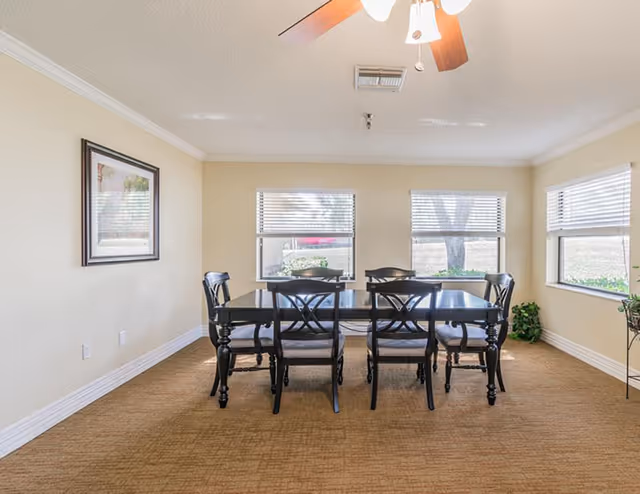 A dining room with a rectangular black dining table and six matching chairs. The room has beige walls, three windows with white blinds, a ceiling fan with wooden blades, a framed picture on the left wall, and a small plant in the corner near the right window.