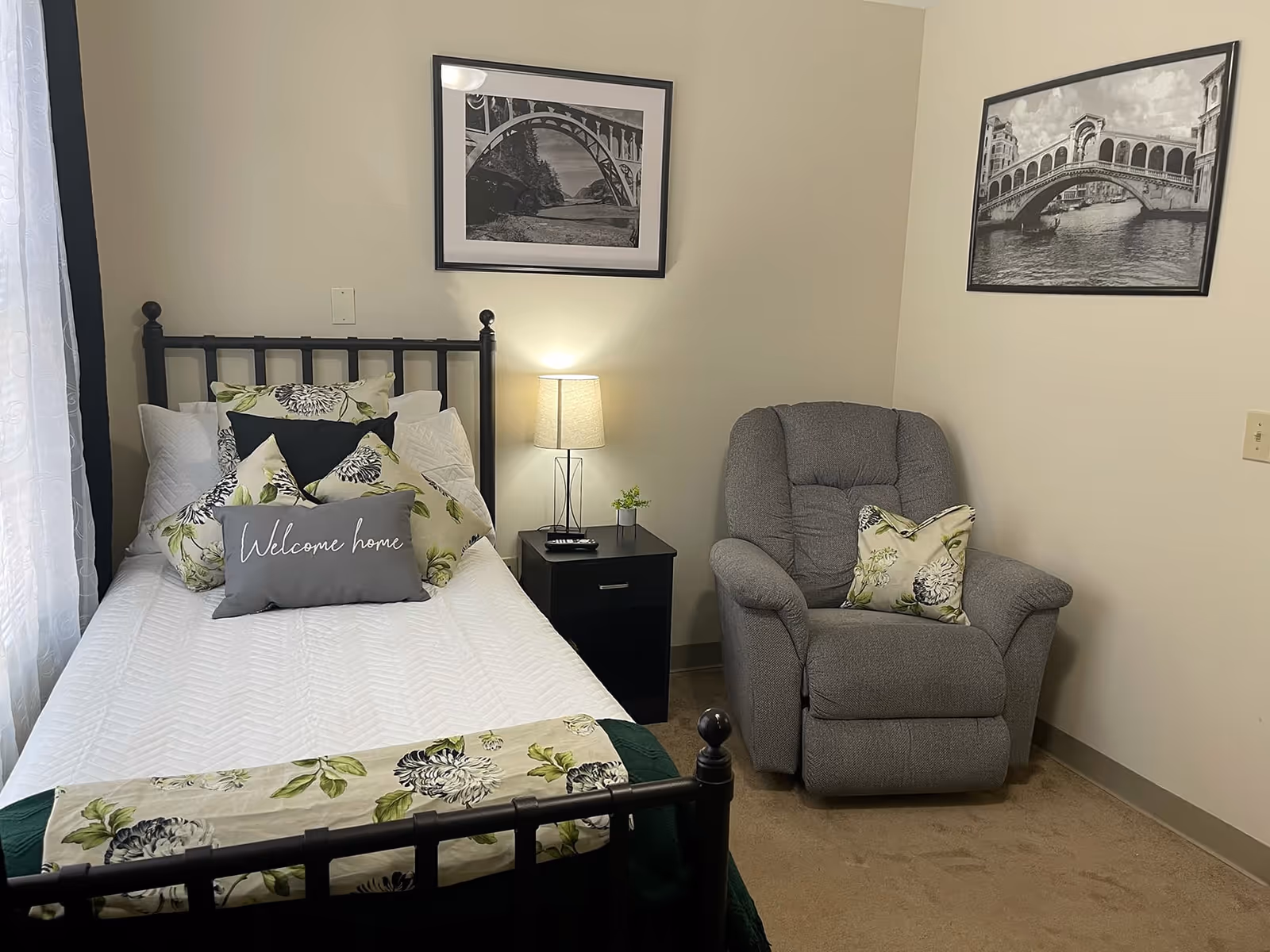A cozy bedroom with a single bed featuring white bedding and multiple floral pillows, including one that says 'Welcome home'. Next to the bed is a black nightstand with a lamp and a small plant. A comfortable gray recliner chair with a floral pillow is positioned in the corner. Two black and white framed pictures hang on the beige walls.