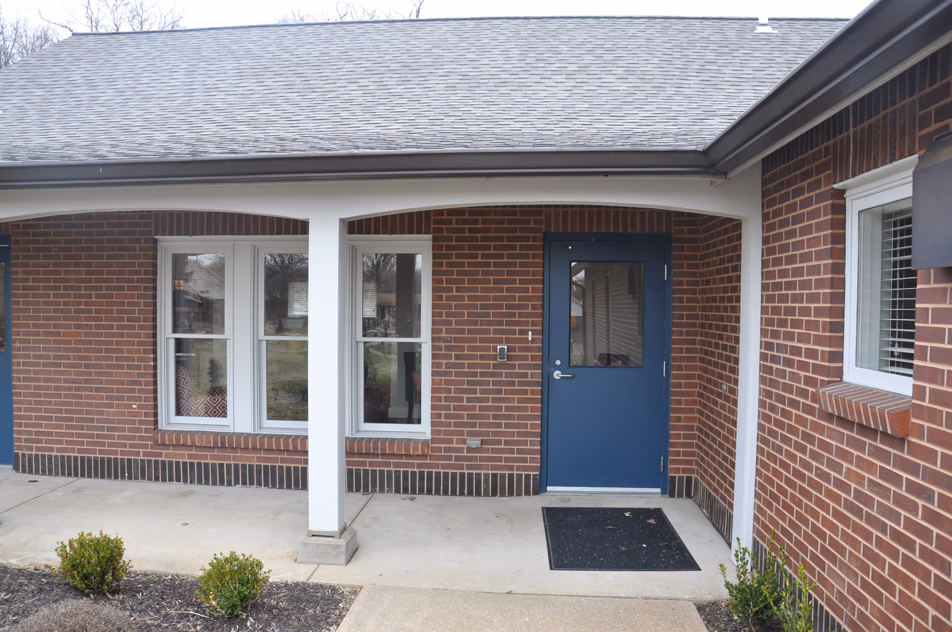Covered entrance of a brick building with a blue door, adjacent windows, and a small concrete porch with shrubs.