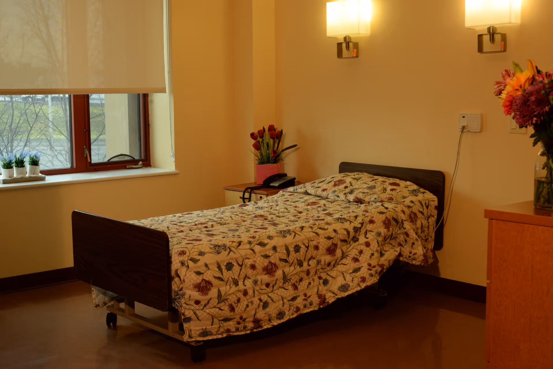 A single hospital-style bed with a floral patterned quilt in a softly lit room. The bed is positioned near a window with a beige roller shade partially down. There are small potted plants on the windowsill, a nightstand with a telephone and a vase of flowers, and a wooden dresser with another vase of flowers. Two wall-mounted lights illuminate the room.