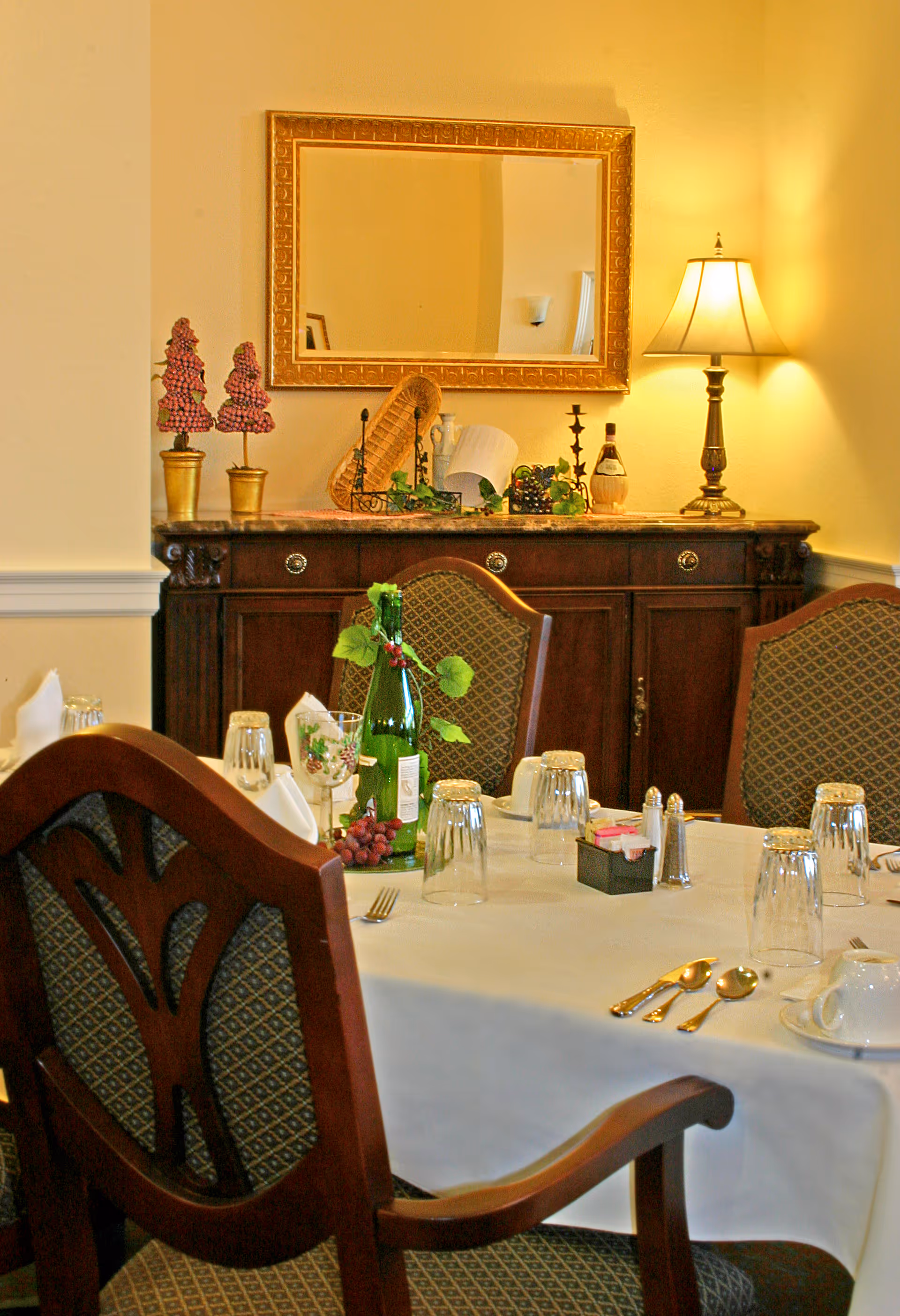A dining room table set with a white tablecloth, glassware, silverware, napkins, and a green bottle decorated with grapevine leaves and grapes. Behind the table is a wooden sideboard with decorative items including a lamp, a mirror with a gold frame, and ornamental grape clusters in gold pots. The chairs around the table have patterned upholstery and wooden frames.