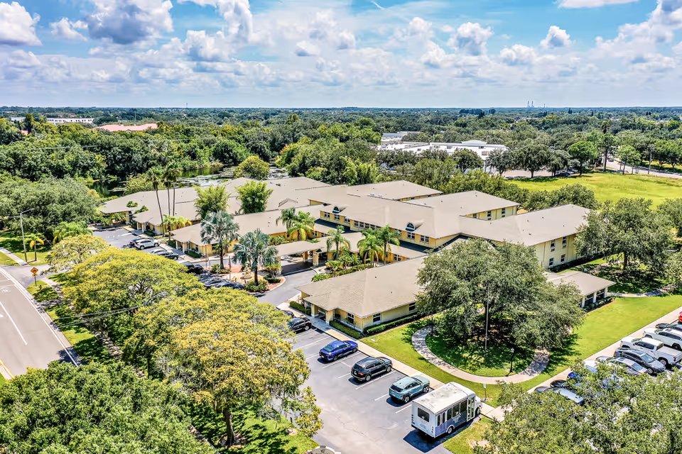 Aerial view of Sun City Senior Living facility surrounded by lush green trees and landscaping. The building has a beige roof and is situated near a road with parked cars and a shuttle bus in the parking lot. The sky is partly cloudy with blue patches.