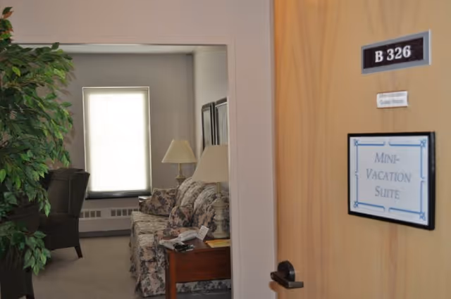 View through an open door labeled B 326 Mini-Vacation Suite into a cozy living room with a floral patterned sofa, two table lamps, a side table with a phone, a large window with blinds, and a green armchair partially visible next to a tall leafy plant.
