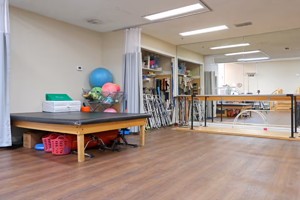 A physical therapy or rehabilitation room with a large padded table, exercise balls, walkers, and various therapy equipment. The room has wooden flooring, a large mirror on one wall, and a ballet barre for support during exercises.