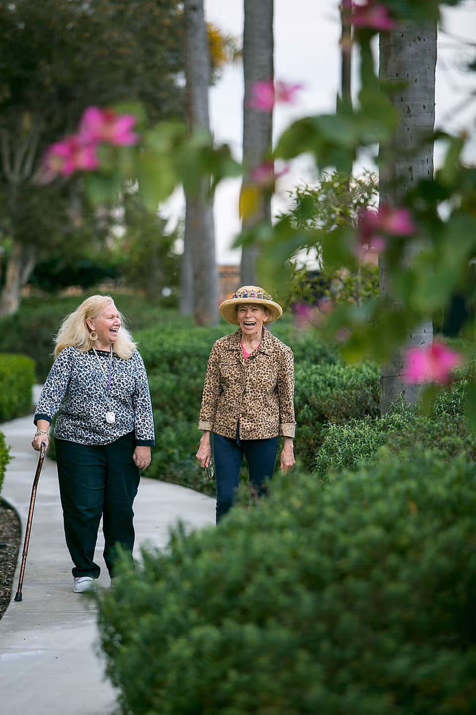 Two elderly women walking and laughing together on a paved garden path surrounded by green bushes and trees. One woman uses a cane and wears a leopard print top, while the other wears a leopard print jacket and a straw hat. Pink flowers are visible in the foreground.