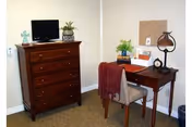 A small room corner featuring a wooden chest of drawers with a small TV and a plant on top, next to a wooden desk with a chair. The desk has a lamp, a clock, a plant, and a corkboard on the wall above it. The floor is carpeted and the walls are light-colored.