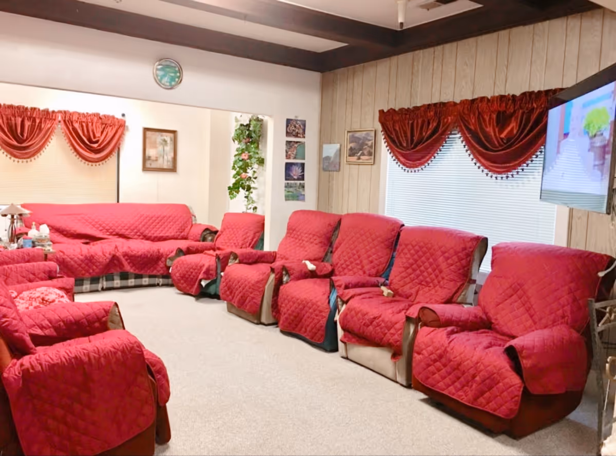 A communal living room with multiple red-quilted-covered recliners and sofas arranged facing a wall-mounted TV.